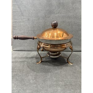 May include: A copper chafing dish with a polished finish, featuring a round lid with a dark brown wooden knob handle. The dish has a long, matching dark brown wooden side handle and rests on a curved metal stand with a central burner component.