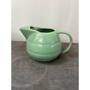 May include: A light green ceramic pitcher with a rounded, ribbed body, a wide pouring spout, and a curved handle. The surface has a smooth, matte finish, and the design features horizontal ridges encircling the main body of the vessel.