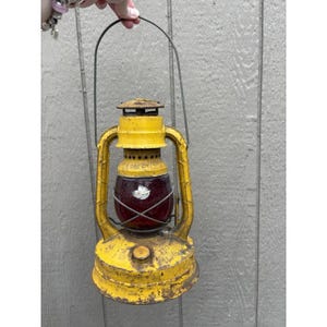 May include: A vintage yellow metal lantern featuring a dark red glass globe protected by a wire cage. The lantern has a weathered, distressed finish with visible signs of rust and wear, a top carrying handle, and a circular base reservoir.