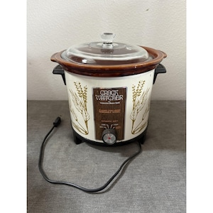 May include: A vintage Crock Watcher by Hamilton Beach slow cooker with a cream-colored exterior featuring gold wheat stalk illustrations. It has a brown removable vessel, a clear glass lid, black side handles, and a front control knob for settings.
