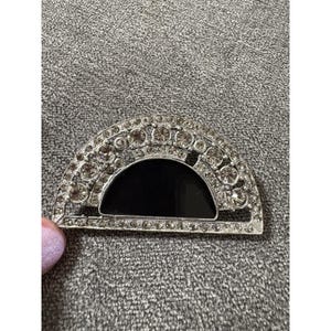 May include: A silver-toned, semi-circular brooch with a black enamel center. The brooch is encrusted with clear rhinestones, creating a sparkling effect. The design is reminiscent of Art Deco jewelry.