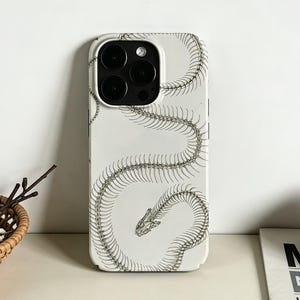 May include: A white phone case featuring a detailed, stylized illustration of a snake skeleton in gray. The design wraps around the case, showcasing the spine and skull. The case has a glossy finish and is designed for a smartphone.