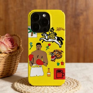 May include: A yellow phone case with a design featuring a boxer in red gloves, a rider on a horse, and a tiger. The case has a black camera lens area and a woven texture. The design includes a rose, a bottle, and a mug.