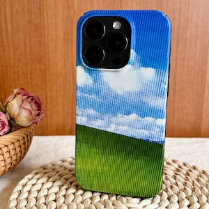Può includere: Una custodia per telefono blu e verde con un design paesaggistico. La custodia raffigura un cielo blu con nuvole bianche e un campo verde. La custodia del telefono ha un modulo fotocamera nero.