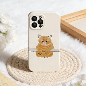 May include: A cream-colored phone case featuring a watercolor illustration of an orange tabby cat. The cat is depicted sitting and looking forward. The case has a camera cutout and is on a woven coaster.