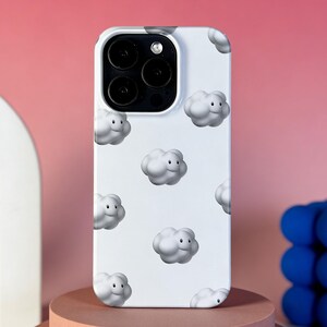Pode incluir: Capa de telefone branca com um padrão repetido de nuvens sorridentes renderizadas em 3D. A capa tem um recorte para a câmera e é exibida em uma superfície rosa com um objeto decorativo azul ao fundo. Capa para smartphone.