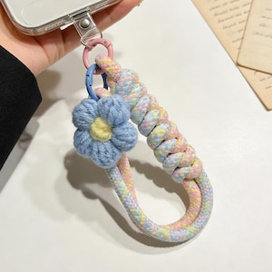 Puede incluir: Un charm de teléfono de color pastel con un acento de flor azul. El charm presenta un cordón trenzado en tonos rosa, amarillo y azul, conectado a una funda transparente para teléfono. Una flor azul con un centro amarillo está unida al cordón.