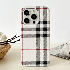 May include: A white phone case with a plaid pattern in black, red, and gray. The case has a camera cutout and is designed to fit a smartphone. The case is on a white surface.