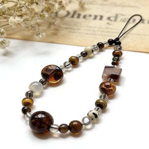 May include: A beaded phone charm featuring a variety of brown, clear, and cream-colored beads. The charm includes round, square, and oval-shaped beads, with a black cord loop for attachment. The beads are strung together with clear string.