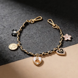 Puede incluir: Una pulsera dorada con eslabones de cadena negros y varios dijes. Los dijes incluyen una moneda, un lazo negro, una flor rosa y dos colgantes en forma de corazón, uno con un osito de peluche y otro con un lazo.