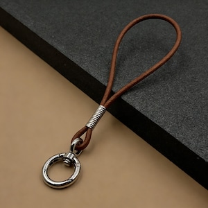 Pode incluir: Um porta-chaves de couro castanho com um fecho de metal prateado e uma argola. O porta-chaves tem um design simples, com uma correia de couro e um anel de metal para prender as chaves. O anel de metal tem aproximadamente 2.5 cm de diâmetro.