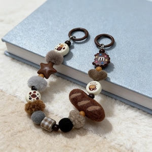 May include: A whimsical keychain featuring various charms, including a star, heart, and bread-shaped felt piece. The keychain also includes decorative pom-poms and round charms with food-related designs. The keychains are attached to brown metal rings.