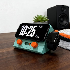 Puede incluir: Una estación de carga de color turquesa con perillas naranjas, que sostiene un teléfono inteligente negro que muestra la hora 10:25. Un Apple Watch negro descansa a un lado. Una pequeña planta suculenta y un altavoz en el fondo.
