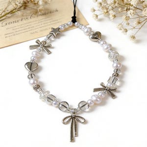 May include: A delicate beaded phone charm featuring a mix of white pearls, clear beads, and silver-toned charms. The charms include hearts, stars, bows, and a rose. The charm is attached to a black cord.