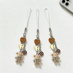 May include: Three phone charms with white straps, silver chains, and decorative elements. Each charm features a small teddy bear, brown heart-shaped beads, yellow leaf-shaped beads, and checkered round beads. The background is a plain white surface.