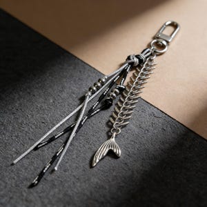 May include: A silver keychain with a mermaid tail charm and a decorative leaf design. The keychain features black, white, and grey cords, along with silver beads and a clasp.
