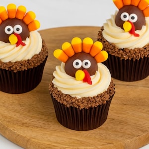 May include: Three Thanksgiving-themed cupcakes on a wooden surface. Each cupcake has a brown chocolate base, vanilla frosting, and is decorated with a turkey made of candy. The turkeys have candy corn feathers, chocolate heads, and candy eyes and beaks.