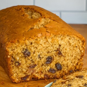 May include: A loaf of golden-brown raisin bread, freshly baked. The bread has a slightly cracked top and is studded with dark raisins. A slice of the bread is cut and visible on the right side of the image, showing the interior texture.