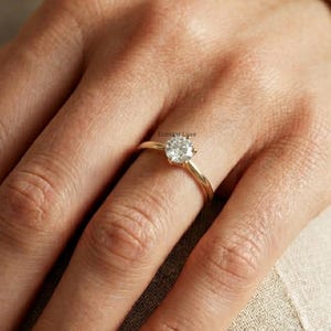 May include: A gold ring with a round, clear gemstone. The ring is on a finger, with the hand and arm visible. The ring's band is thin and yellow. The background is a neutral color.