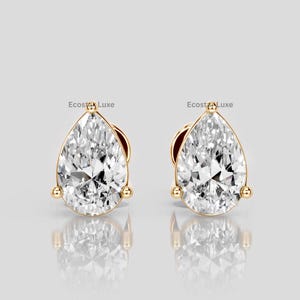 Puede incluir: Un par de pendientes de oro, cada uno con una piedra preciosa clara en forma de pera. Los pendientes tienen un engaste de oro con pequeños detalles redondos. La marca "Ecosta Luxe" es visible en cada pendiente.