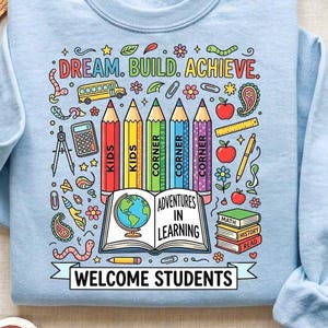 Puede incluir: Una sudadera azul claro con un gráfico colorido que incluye el texto "DREAM. BUILD. ACHIEVE.", "ADVENTURES IN LEARNING" y "WELCOME STUDENTS". El diseño muestra cinco lápices, un libro abierto con un globo terráqueo, un autobús escolar y herramientas matemáticas.