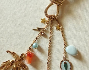Puede incluir: Llavero dorado con múltiples colgantes. Los colgantes incluyen una flor, un rayo, estrellas, una cuenta azul, un colgante rojo y naranja, un colgante rectangular con un ojo y un colgante ovalado con un pez.