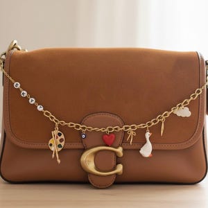 Puede incluir: Un bolso de hombro de cuero marr&oacute;n con una cadena dorada y abalorios decorativos. Los abalorios incluyen una paleta, un coraz&oacute;n rojo, un ganso y una nube. El bolso tiene un gran cierre "C" dorado.