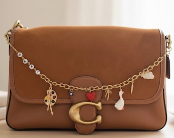 May include: A brown leather shoulder bag with a gold chain and decorative charms. The charms include a palette, a red heart, a goose, and a cloud. The bag has a large gold "C" clasp.