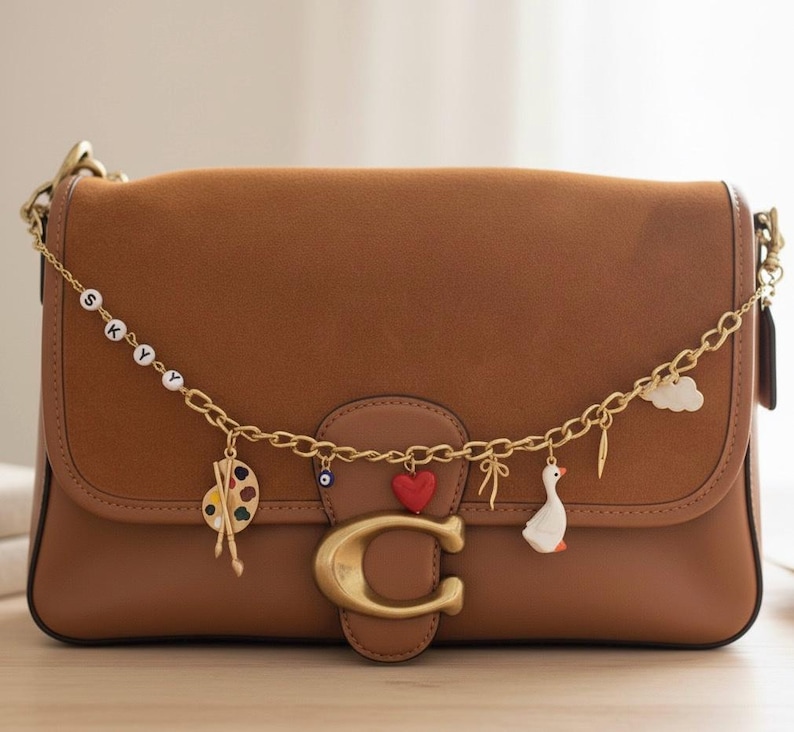 Puede incluir: Un bolso de hombro de cuero marr&oacute;n con una cadena dorada y abalorios decorativos. Los abalorios incluyen una paleta, un coraz&oacute;n rojo, un ganso y una nube. El bolso tiene un gran cierre "C" dorado.