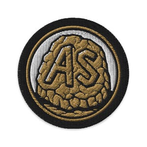 May include: Embroidered patch with a circular design. The patch features the letters "AS" in a textured gold color, set against a white background, and framed by a black border.