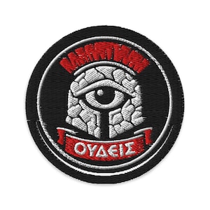 May include: Embroidered patch with a black circular background. The patch features a white eye design with a red banner at the bottom that reads "OYAEIΣ". The top of the patch has a red design.