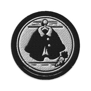 May include: Embroidered patch featuring a black coat design within a white and black circular border. The coat has a high collar and three buttons. The patch is likely for clothing or accessories.