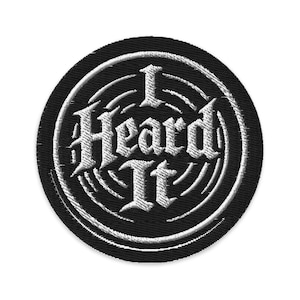Pode incluir: Um patch redondo bordado em preto com a frase "I Heard It" em branco, com letras em estilo gótico. O texto é rodeado por linhas brancas concêntricas, criando um efeito visual. Adequado para casacos, malas ou outros artigos de tecido.
