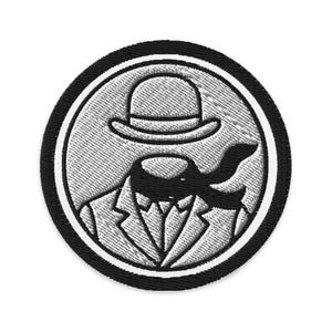 May include: Embroidered patch featuring a stylized illustration of a figure in a suit and bowler hat. The design is primarily black and white, with a textured appearance. The patch has a circular shape with a black border.