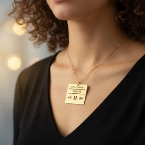 May include: A gold-colored pendant necklace with a square pendant. The pendant features the text "NOW PLAYING" above the name "CHLOE" and music player controls. The necklace is worn against a black top.