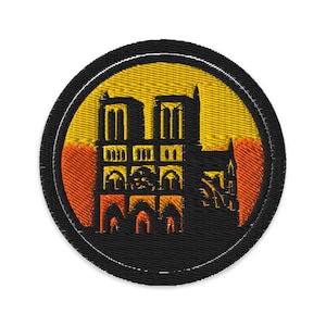 May include: Circular embroidered patch depicting the Notre Dame Cathedral in silhouette. The design features a black outline against a yellow and orange backdrop, framed by a black border. Ideal for customising clothing or accessories.
