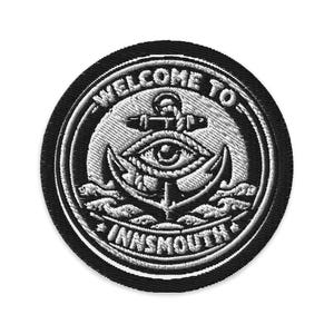 May include: Circular embroidered patch with a black border and a white and grey design. The design features an anchor, an eye, and the words "WELCOME TO INNSMOUTH". The patch is round.
