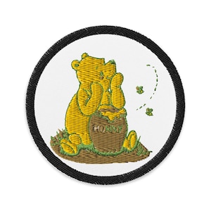 May include: Embroidered patch featuring a yellow bear sitting next to a brown honey pot with the word "HONEY" on it. The bear is on a patch with a white background and a black border. Green grass and three green leaves are also visible.