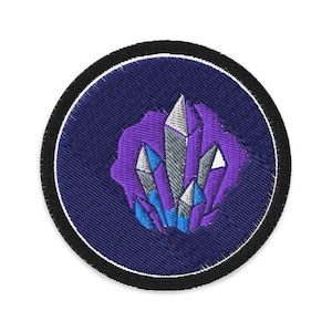 May include: Embroidered patch featuring a cluster of crystals in shades of purple, blue, and silver. The crystals are set against a dark purple background within a circular patch with a black border and white stitching.