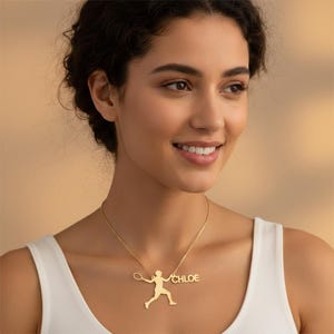 Peut inclure: Un collier doré avec un pendentif représentant un joueur de tennis et le nom "CHLOE". Le pendentif montre un joueur de tennis avec une raquette. Le collier est porté par une personne portant un débardeur blanc.