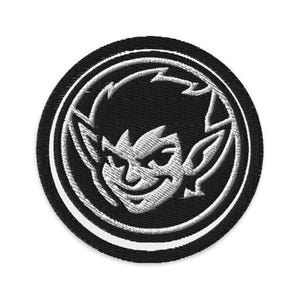 Könnte beinhalten: Gestickter Aufnäher mit einem stilisierten Goblin-Gesicht in Weiß auf schwarzem kreisförmigen Hintergrund. Der Goblin hat spitze Ohren und ein schelmisches Grinsen. Der Aufnäher hat einen weißen Rand und einen schwarzen Außenrand.