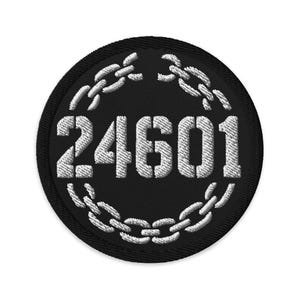 May include: A round, black embroidered patch featuring the number "24601" in white, encircled by a white chain design. The patch is likely for clothing or accessories.
