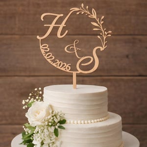 Puede incluir: Un adorno de pastel de madera con las letras "H & S" y la fecha "02.02.2026". El adorno está colocado sobre un pastel blanco de varios niveles, adornado con flores blancas y vegetación. El fondo es una pared de madera.