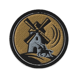 Puede incluir: Parche bordado con un diseño de molino de viento. El parche tiene un borde negro, un fondo dorado y una imagen detallada de un molino de viento en tonos grises y marrones. El molino de viento está sobre un fondo texturizado.