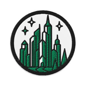 May include: Embroidered patch featuring a stylized cityscape in shades of green and white, encircled by a black border. The design includes buildings and four star-shaped accents, suitable for jackets, bags, or other fabric items.