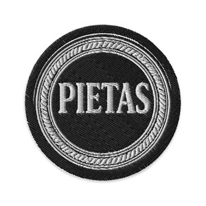 May include: A round, black embroidered patch with the word "PIETAS" in white, centered within a white rope-like border. The patch has a textured appearance, suggesting a fabric material.
