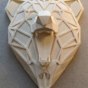 May include: A geometric paper sculpture of a bear's head, with its mouth open, revealing teeth. The artwork is made of off-white paper, with a detailed, angular design. The bear's head is a striking piece of wall decor.