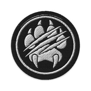 May include: Embroidered patch featuring a white paw print with claw marks inside a white circle, set against a black background. The patch is round and has a stitched border, suitable for jackets, bags, or other fabric items.