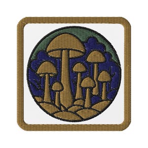 Peut inclure: Écusson brodé représentant un groupe de champignons stylisés dans des tons marron, beige et or, sur un fond bleu et vert. L'écusson a une bordure blanche et un bord cousu marron.