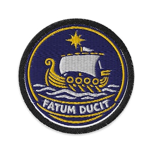 May include: Embroidered patch with a Viking ship design. The ship has a white sail and gold hull, sailing on blue waves. A gold star is above the ship. The patch has a navy blue background with the words "FATUM DUCIT" in white.